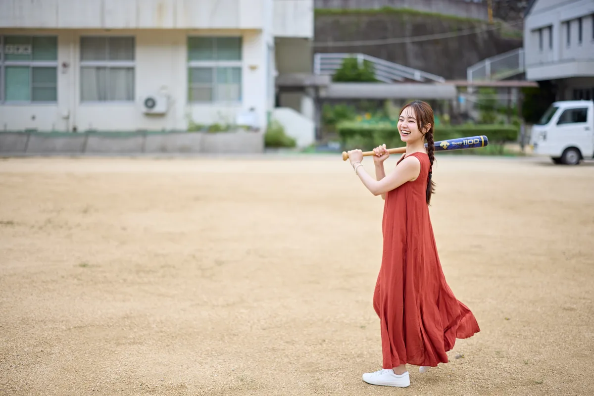 校庭でバットを肩にかついで笑う赤いワンピースの女性