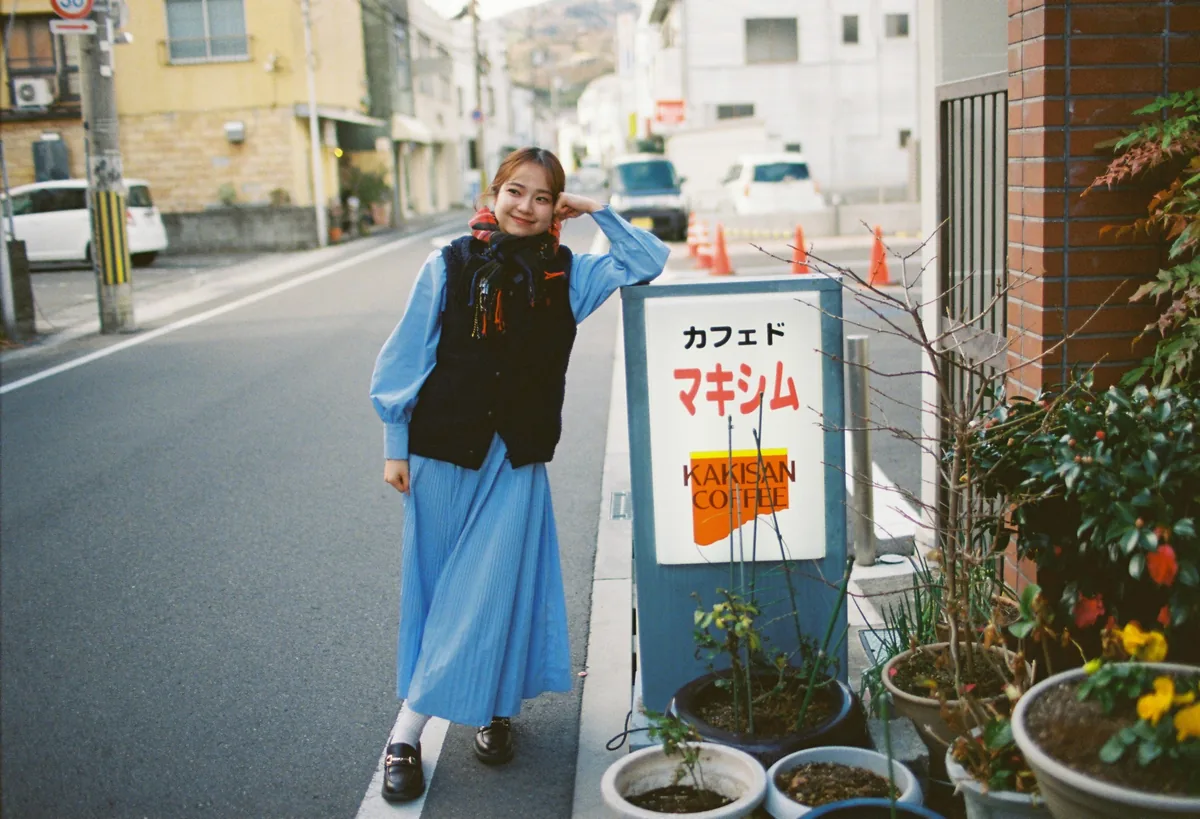 喫茶店の看板にひじをついて立つ、青いワンピースの女性