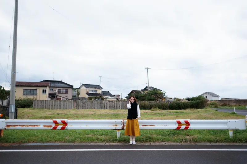 ガードレールの向こう、家並みを背に立つ女性