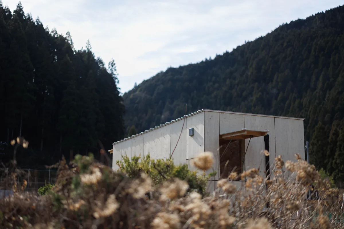 枯れ草の向こう、山を背に建つ木の外壁のモバイルハウス「住箱」