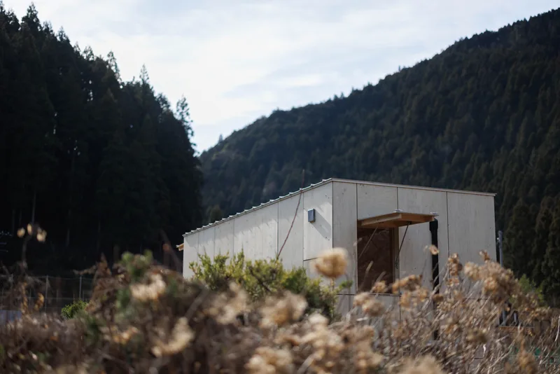 枯れ草の向こう、山を背に建つ木の外壁のモバイルハウス「住箱」